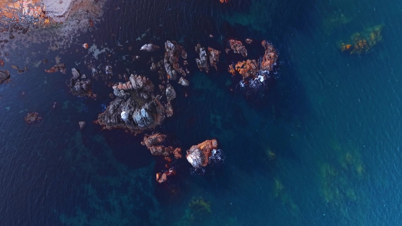 Aerial top down view of rocky outcrops in deep blue sea water. From directly overhead the drone captures red-brown jagged rocks emerging from dark blue water above a shallow reef.