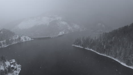Aerial-view-of-a-lake-and-dam-surrounded-by-a-captivating-blend-of-fog-and-snow,-mountain-forest,-in-Dragan-dam-from-Transylvania,-Romania