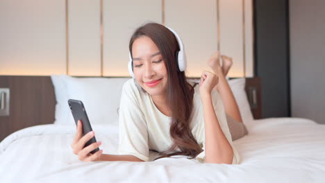 a close-up of a young woman with headphones on and holding a smartphone lays on the bed and rocks out to the music