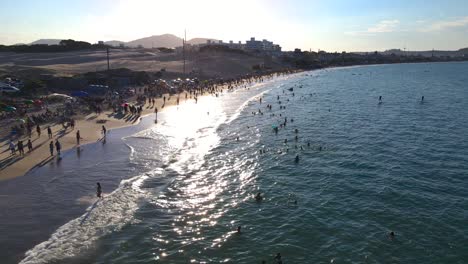 Aerial-scene-of-drone-low-flight-on-the-beach-with-many-people-in-the-sea-and-on-the-sand-tropical-beach-in-sunset-with-sun-in-ocean-summer-in-florianopolis-Ingleses-beach-blue-sky-full-umbrella