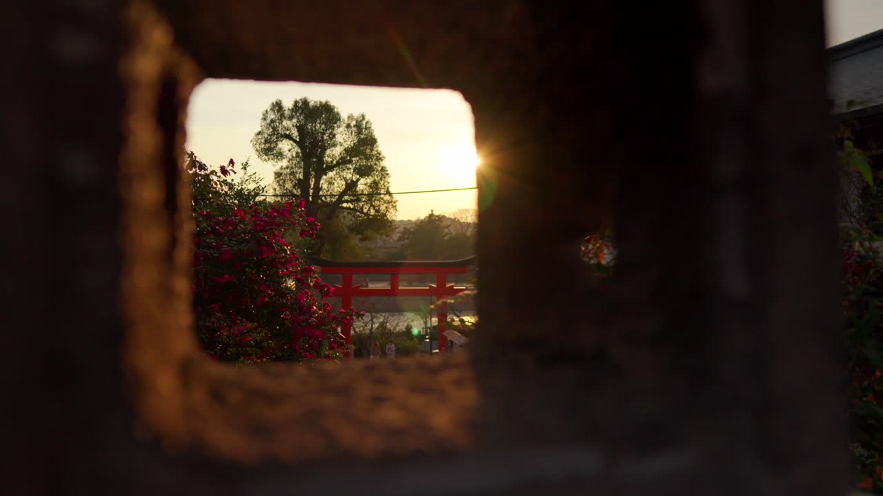 Panorerende udsigt ved solnedgang gennem en traditionel japansk stenlanterne, der afslører den naturskønne Uji-flod og en levende rød torii-port ved Uji-helligdommen i Uji, Japan