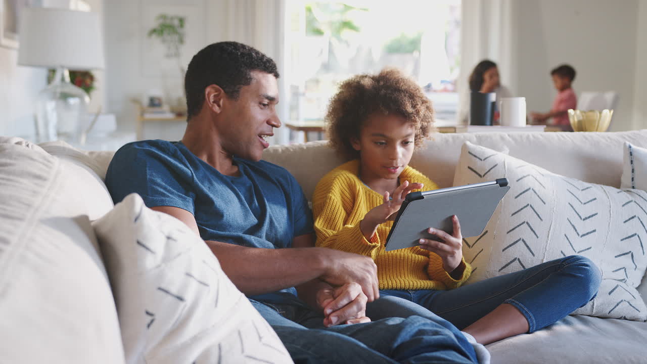 Father and his pre-teen daughter on sofa using tablet computer, mum and toddler in the background