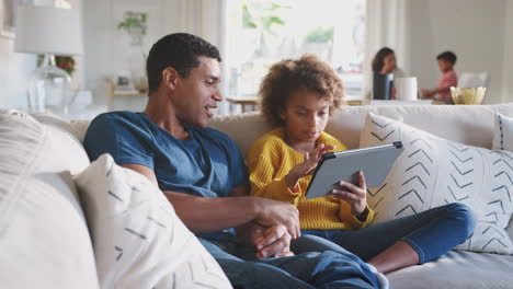 father and his pre-teen daughter on sofa using tablet computer, mum and toddler in the background