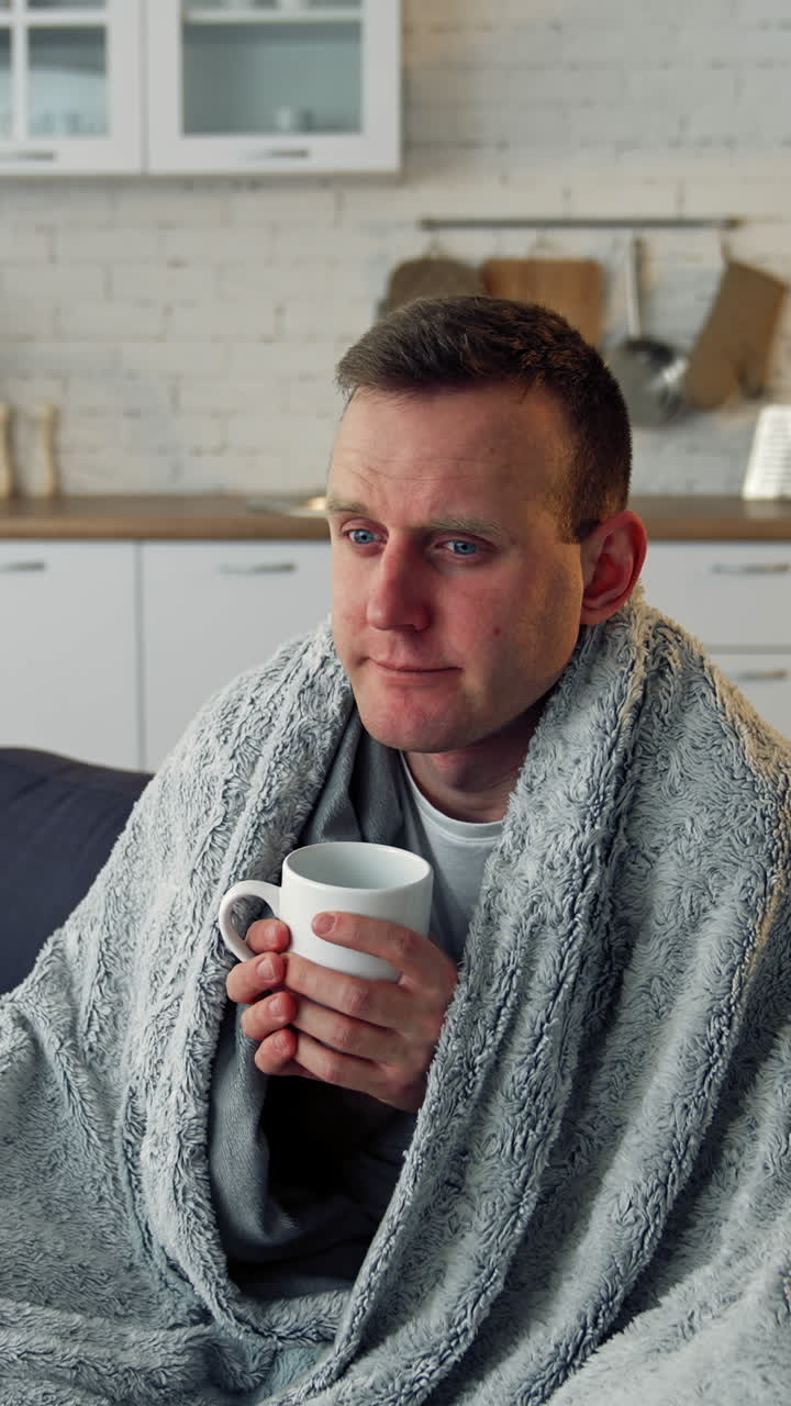 Folk medicine. Sick young man with fever wrapped in warm blanket drinking hot tea. Pandemic. Vertical video