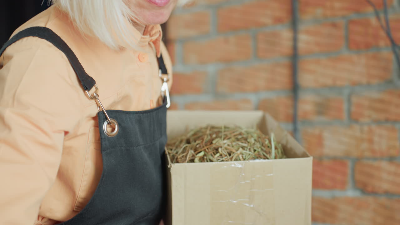 Florist with blonde hair wearing apron working in workshop, close up portrait with thoughtful focused expression, preparing handmade arrangement while standing near brick wall background in creative studio