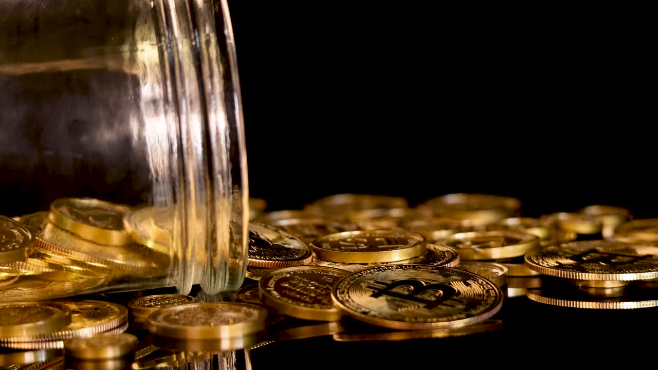Glass jar rotates, releasing gold coins onto reflective black surface under dramatic studio lighting