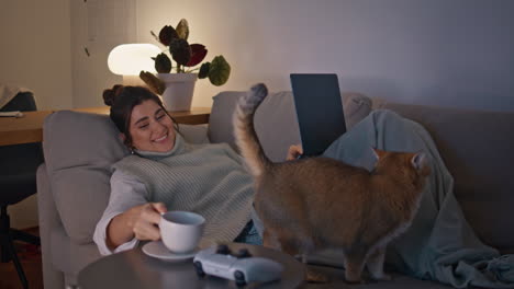 domestic brunette drinking coffee cup lying sofa with laptop at evening room