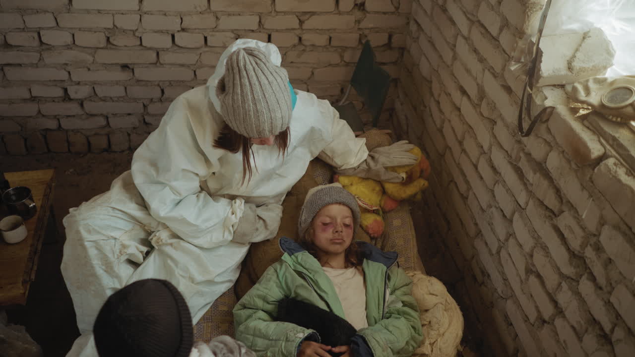 Aerial view of worried survivors in white suits inside brick shelter looking down at unconscious child with bruised eyes lying on mattress beside worn plush toy and blanket, suggesting illness