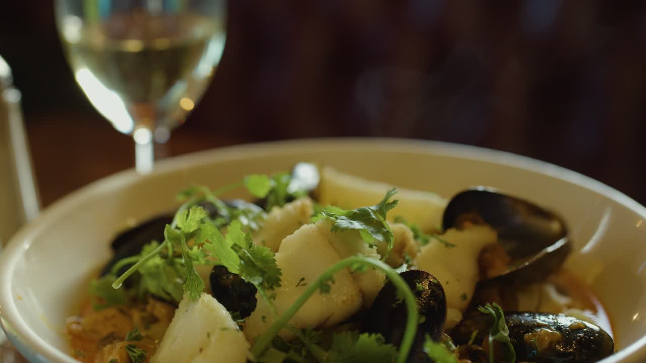 A hand uses a spoon to serve steaming seafood chowder with mussels, scallops, and herbs in a warmly lit restaurant setting, with wine glass visible