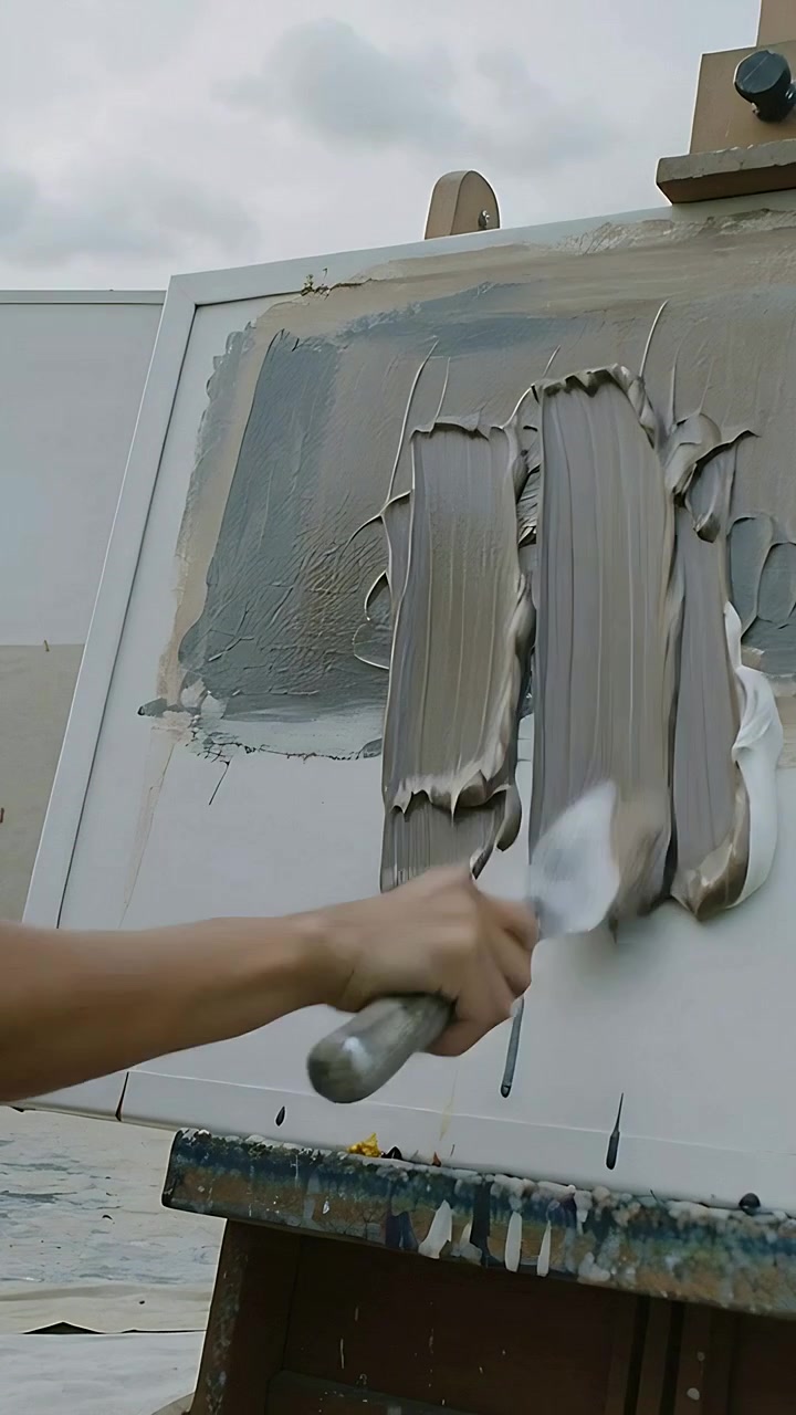 Vertical video: Applying knife artist in grey tee pulling paint on canvas by wall