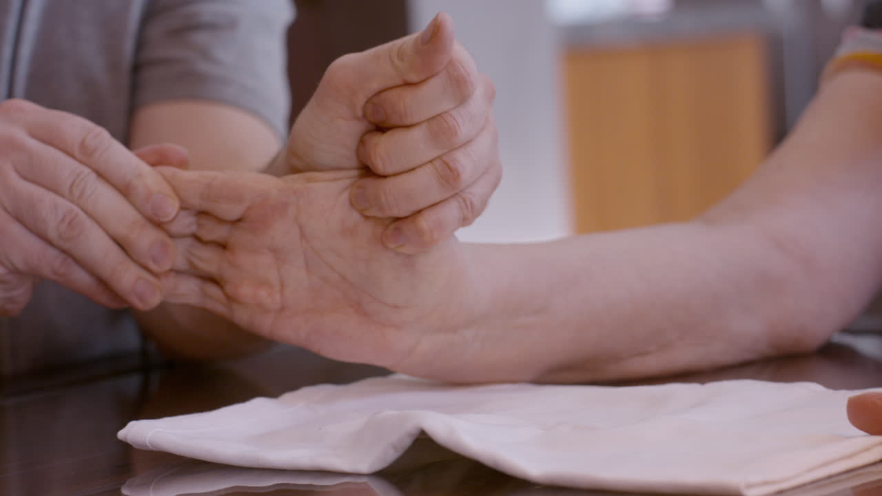 A male caregiver assists a senior female stroke survivor by gently stimulating and mobilizing her hemiplegic right thumb after stretching her hand; at the kitchen table, on cloth pad.