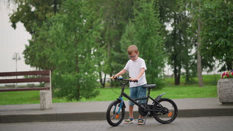 a young boy wearing glasses holds onto a bicycle, carefully steadying it on a paved path, he is dressed in a white top and shorts, with trees and greenery surrounding