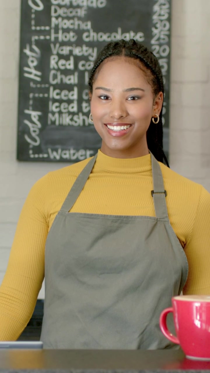 Vertical video: Reaching barista in gray apron placing red mug on cafe counter, serving and smiling