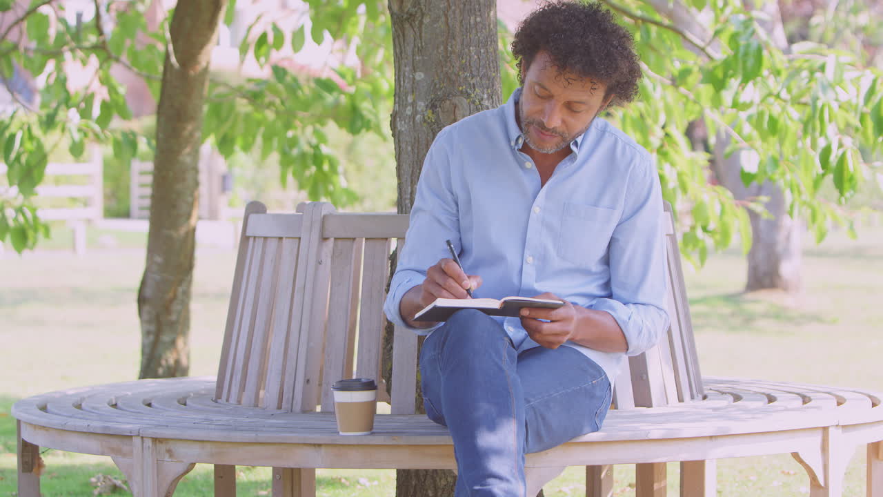 Mature Man Sitting On Park Bench Under Tree Writing In Notebook Or Diary