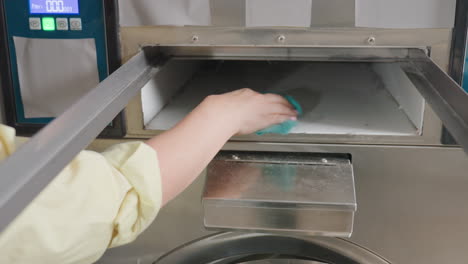 High angle view of laundry owner hands cleaning inner filter drawer of industrial washing machine with hand towel, stainless panel, hygiene maintenance, safe service routine inside laundromat