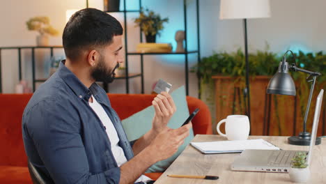 Indian-man-freelancer-using-credit-bank-card-and-smartphone-while-transferring-money-at-home-office