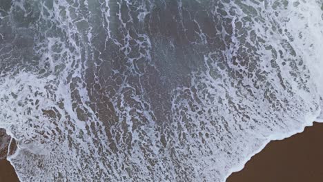 Ocean-waves-crash-and-spread-seafoam-across-dark-brown-sand-swirling-as-water-recedes,-aerial-bird's-eye-view