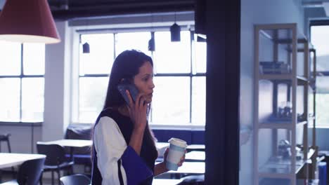 Geschäftsfrau-Mit-Gemischter-Abstammung-Spricht-Auf-Dem-Smartphone-Und-Hält-Kaffee-In-Der-Hand,-Während-Sie-Im-Bürokorridor-Läuft