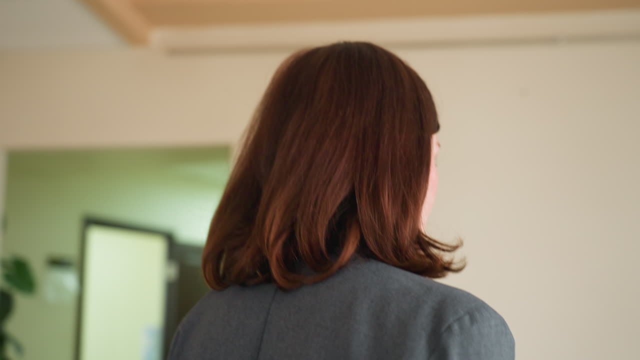 Profile view of businesswoman with auburn hair in gray blazer standing in office hallway. Professional female looking toward corridor with doors visible in background. Corporate setting with cream walls