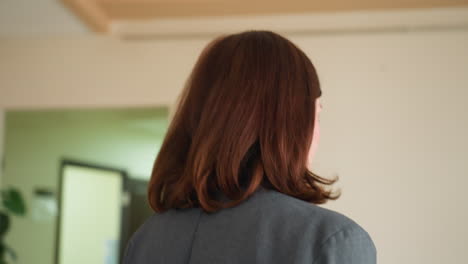 profile view of businesswoman with auburn hair in gray blazer standing in office hallway. professional female looking toward corridor with doors visible in background. corporate setting with cream walls