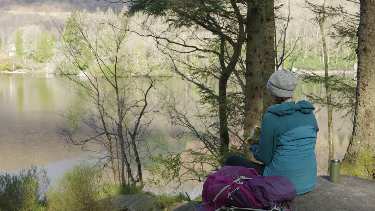 A woman sits on a rock eating her lunch while out for a walk in the forest, looking out across a Scottish Loch and hillside in the sun