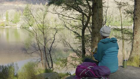 a woman sits on a rock eating her lunch while out for a walk in the forest, looking out across a scottish loch and hillside in the sun