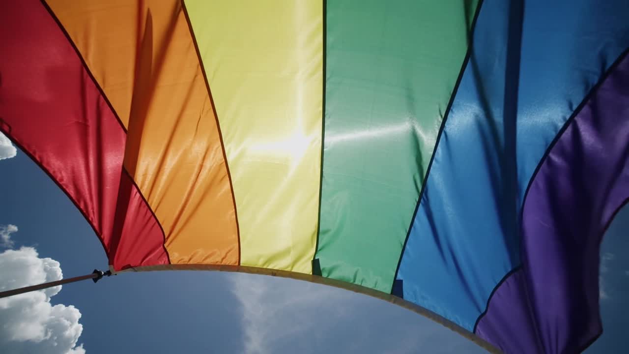 Grande bandiera a strisce arcobaleno che sventola mossa dal vento davanti all'obiettivo all'aperto, riflettendo i raggi del sole.