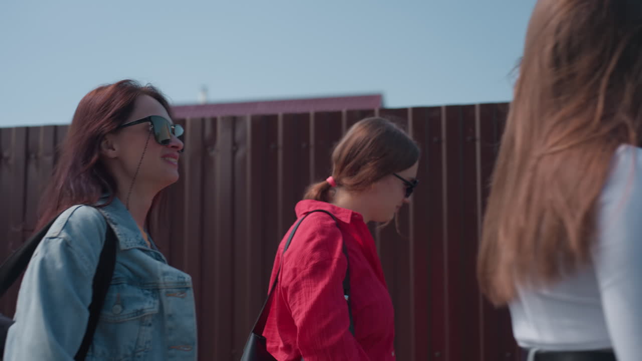 Three cheerful girls walk beside wooden fence under sunny sky, laughing and talking with relaxed expressions, one adjusting hair while others raise hands mid-conversation in warm afternoon light