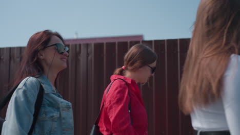 three cheerful girls walk beside wooden fence under sunny sky, laughing and talking with relaxed expressions, one adjusting hair while others raise hands mid-conversation in warm afternoon light