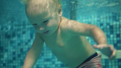 cute blonde toddler is diving under the water in the swimming pool and swimming there until his mother helps him to get out. and