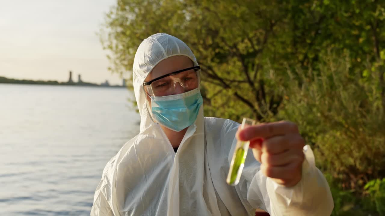 Operaio con mascherina raccoglie un campione d'acqua accanto al fogliame verde in riva al lago, luce soffusa e spazio aperto