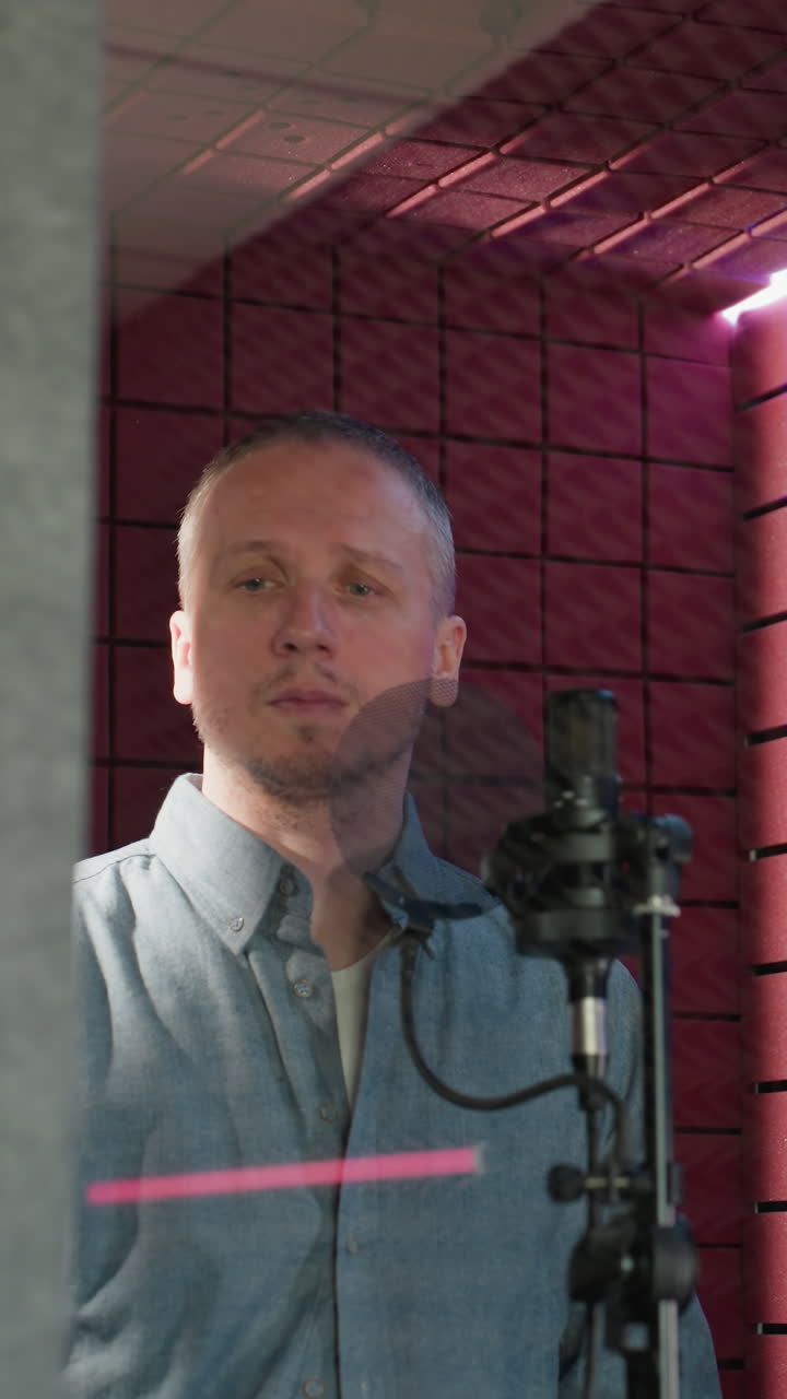 un hombre con una camisa de manga larga azul se encuentra frente a un micrófono de estudio en una habitación a prueba de sonido roja. la escena captura un momento en un entorno de grabación profesional