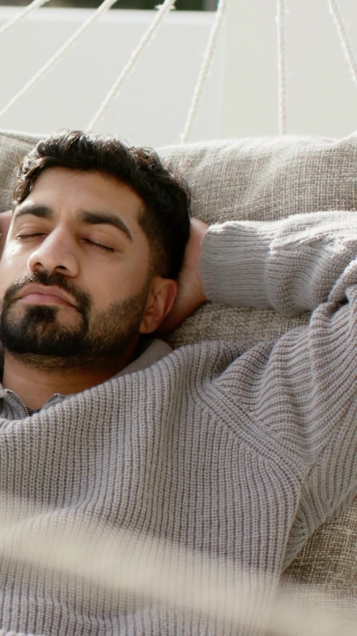 Video verticale: Riprese di un uomo che si rilassa su un'amaca a casa per riposare, indossando un maglione grigio su un cuscino.