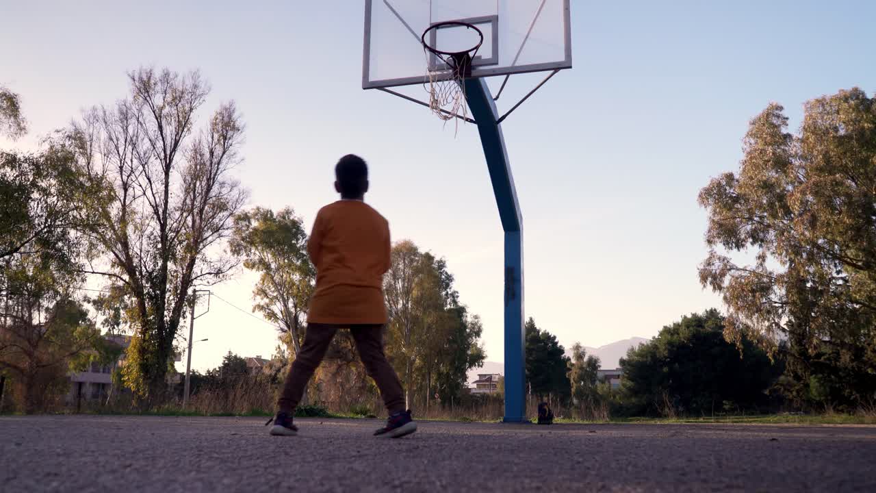 nivel del suelo, vista baja del niño caucásico que intenta tiros de baloncesto, pierde el balón de aire, en la cancha de baloncesto de espacio abierto, árboles en el fondo
