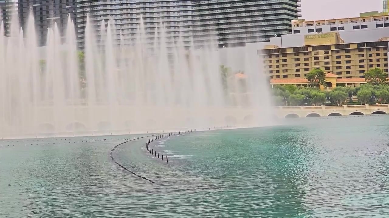 Escena de viaje luminosa con un gran espectáculo de fuentes sobre un lago de resort turquesa, con chorros de agua altos, neblina, arcos de piedra, árboles verdes, edificios hoteleros circundantes y un horizonte soleado