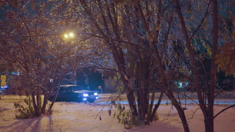 nighttime scene of a car passing by snow-covered trees and illuminated urban streets, with warm streetlights casting golden hues and shadows over the snowy ground