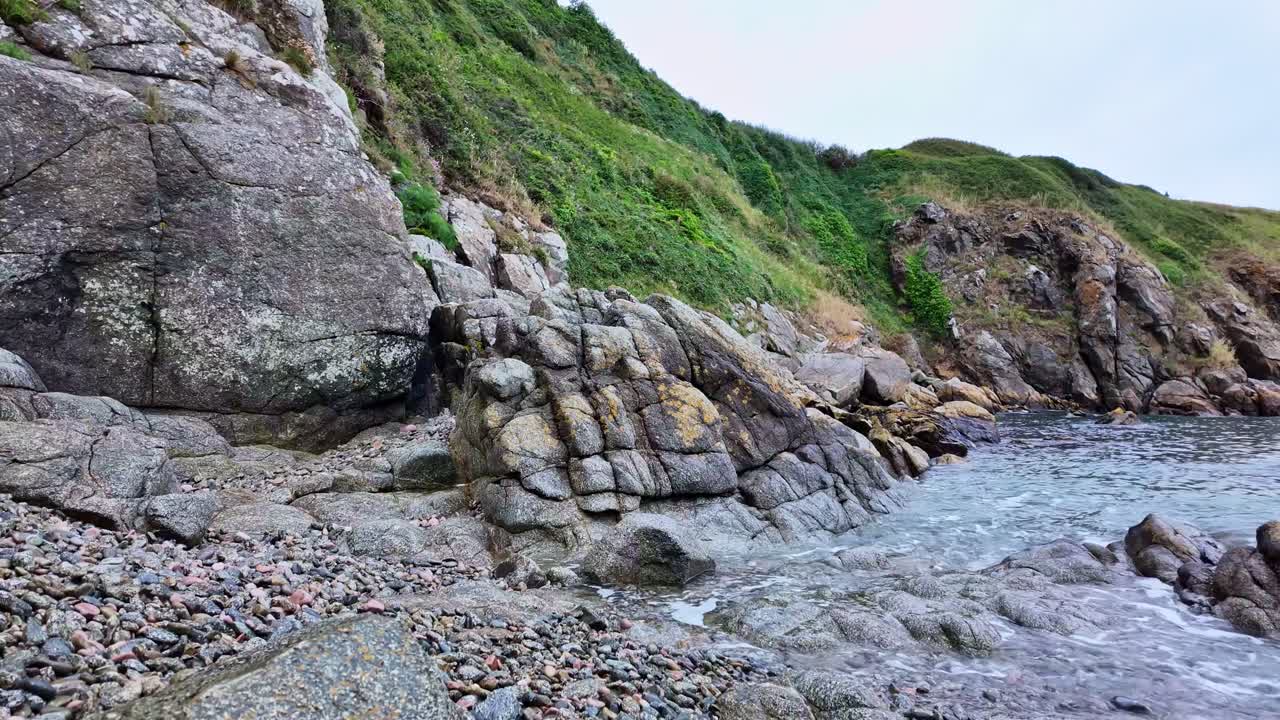 The camera moves downward along a green cliff to reveal waves crashing against rocks by the sea. Coastal scene with vegetation and water -Brittany France