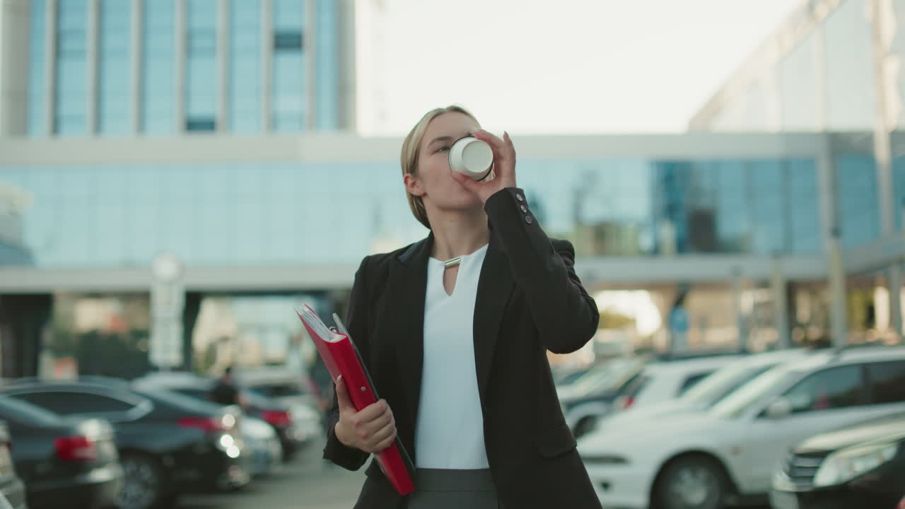 White lady in professional outfit walking along path of parked cars holding red folder and drinking beverage from takeaway cup with glass residential building and modern cityscape in background