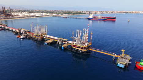 Drone shot panning horizontally along offshore oil station with moored ship, refinery connection, storage tanks, and Sicilian bay in background