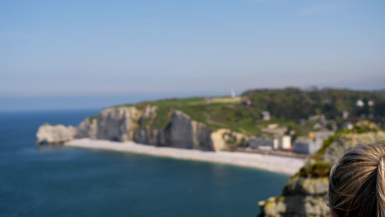 primer plano de una mujer rubia disfrutando de la vista de la ciudad de etretat con acantilados marinos durante un día ventoso - cambio de enfoque