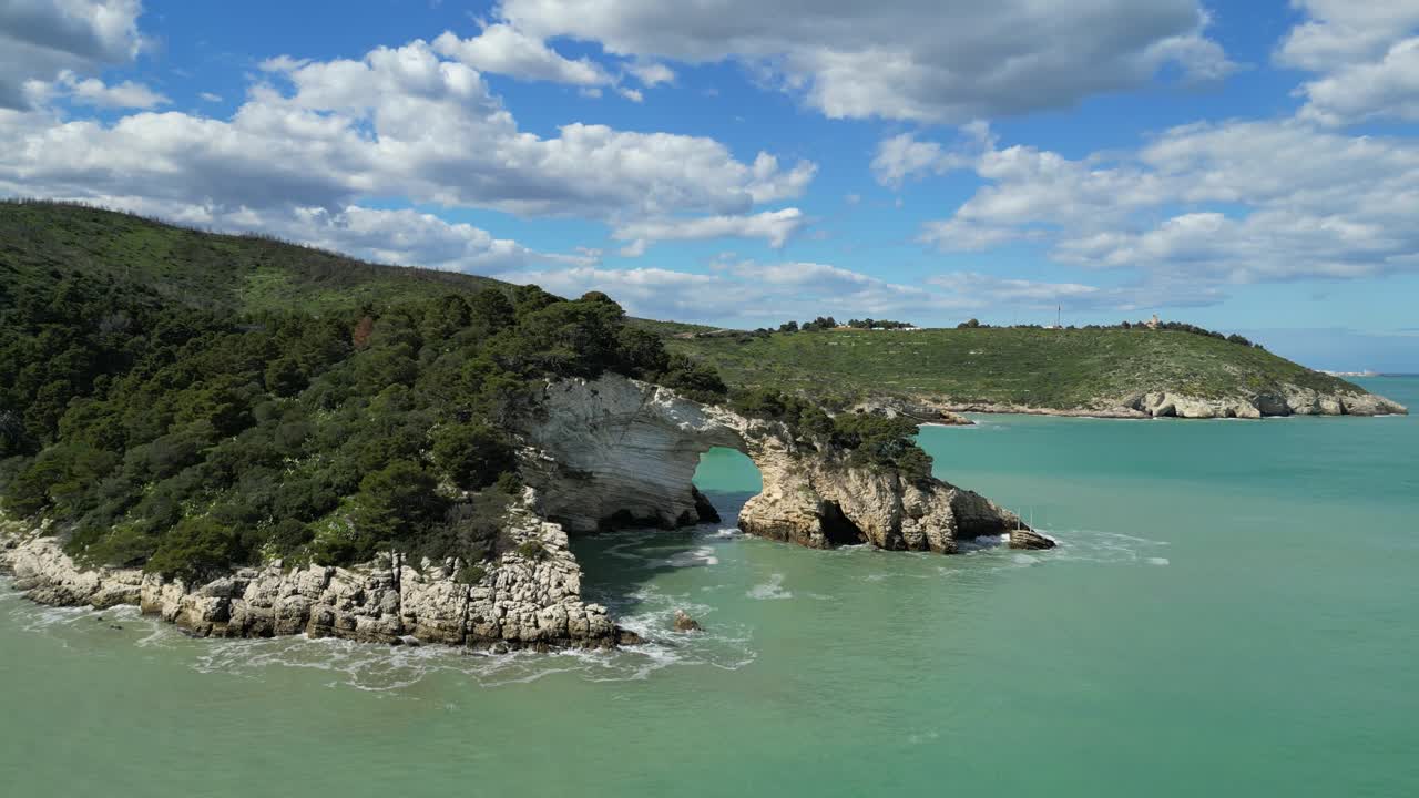 Stunning 4K drone footage of a natural sea arch, rugged limestone cliffs, turquoise waters, lush coastal vegetation along the Gargano Peninsula in Puglia, southern Italy