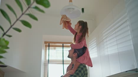 una pequeña chica morena en un vestido rosa se sienta en los hombros de su padre moreno en una camisa a cuadros verdes y limpia la lámpara en el techo con un paño especial en una cocina moderna