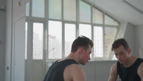 teenager in black vest and pants walks to chair by large mirror in bright studio while open case with security gear rests on floor near window letting in soft natural light and urban skyline