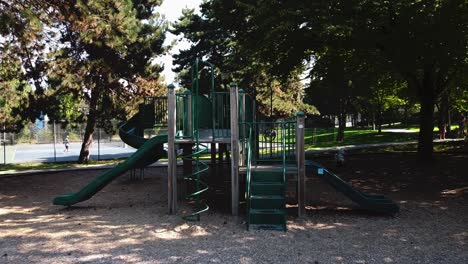 Leerer-Spielplatz-Mit-Schildern,-Die-Kinder-Verbieten,-Und-Lockdown-Covid-19-Beschränkungen-Der-Sozialen-Distanzierungspandemie-In-Einem-üppig-Grünen-Sommerpark-Mit-Aktiven-Menschen,-Die-Im-Hintergrund-Tennis-Spielen-2-2