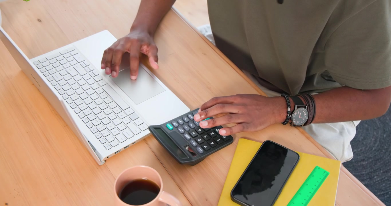 Studente maschio che digita su un laptop bianco, spostando la mano destra su una calcolatrice nera, verificando i numeri