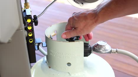a hand turns a valve on a gas grill tank outdoors. bright lighting highlights the action in a casual setting