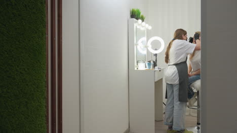beautician works on blond client seated on furry chair in well lit salon with white walls and green accent wall, featuring mirror setup with ring light and cosmetic tools neatly arranged on table