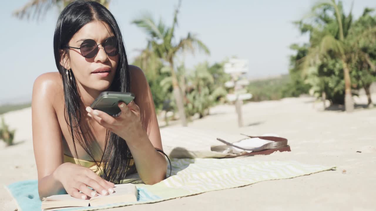 Donna ispanica felice con occhiali da sole sdraiata in spiaggia che parla allo smartphone, spazio per il testo, slow motion