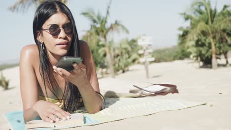 happy hispanic woman in sunglasses lying on beach talking on smartphone, copy space, slow motion