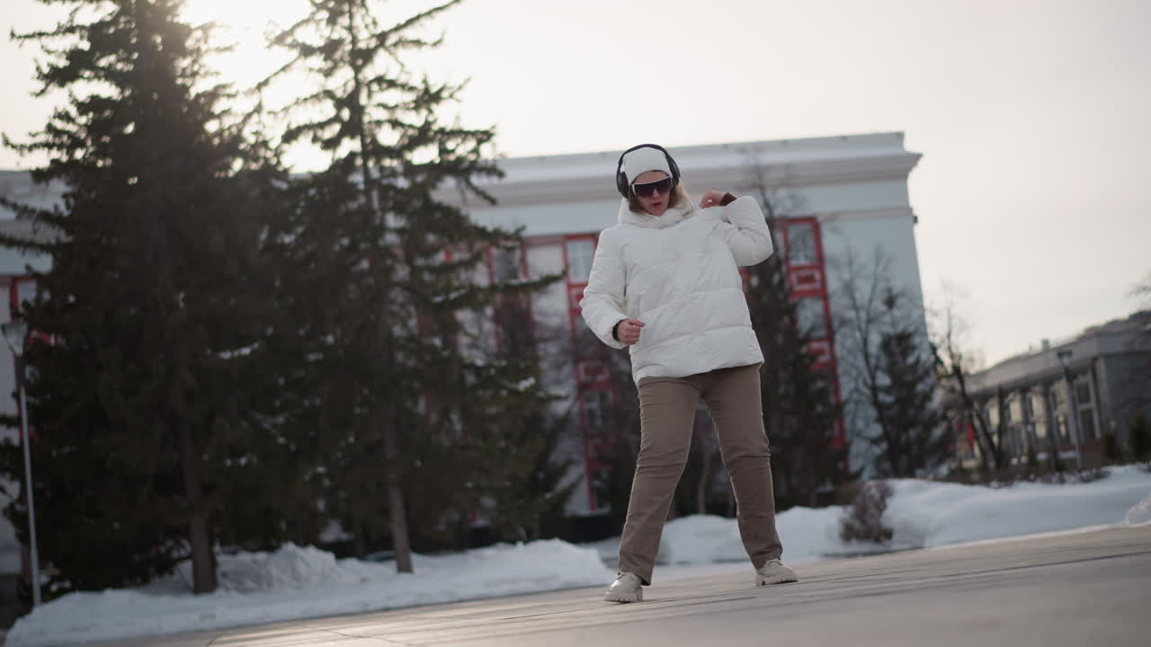 Student dancing excitedly rolling hands moving feet smiling and lifting arms in snowy plaza wearing winter coat and knit beanie captured turning around under pale sky with joyful rhythmic energy